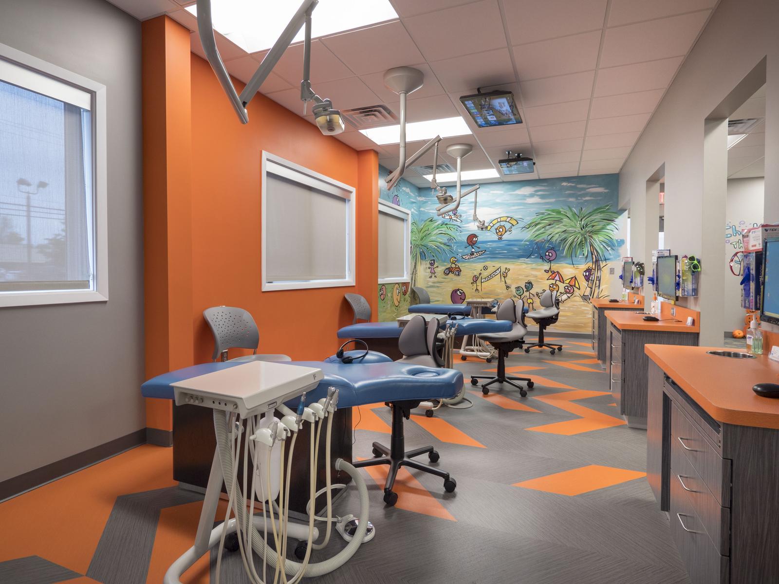 Wide view of the open bay treatment area showing several dental chairs, orange cabinetry, and the beach-themed mural