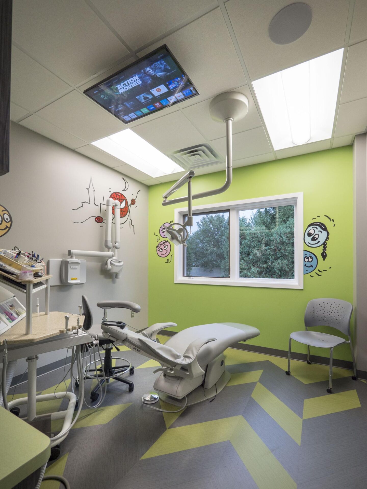 Private pediatric treatment room with a ceiling-mounted TV, superhero wall decals, and a dental chair facing a window
